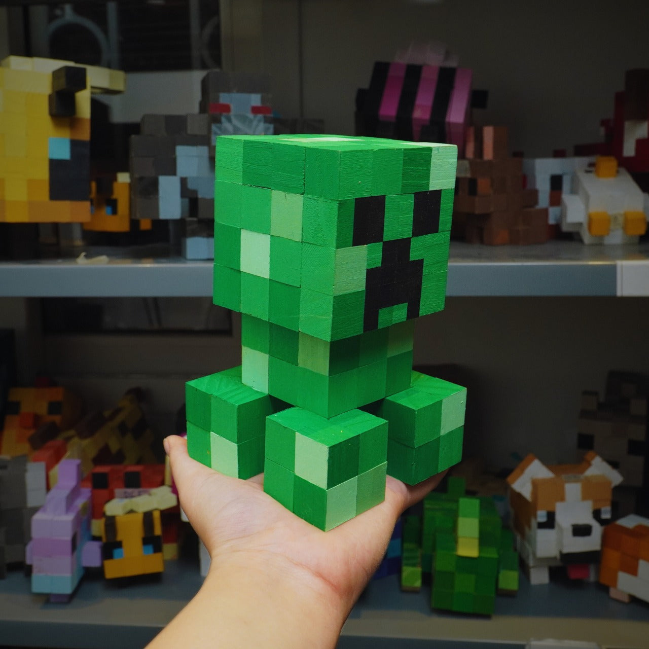 Wooden Minecraft Creeper handmade figure displayed on hand with pixel toy shelf background.